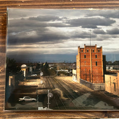 Vintage photograph of a cityscape with a tall brick building and train tracks, framed in glass on a wooden surface.