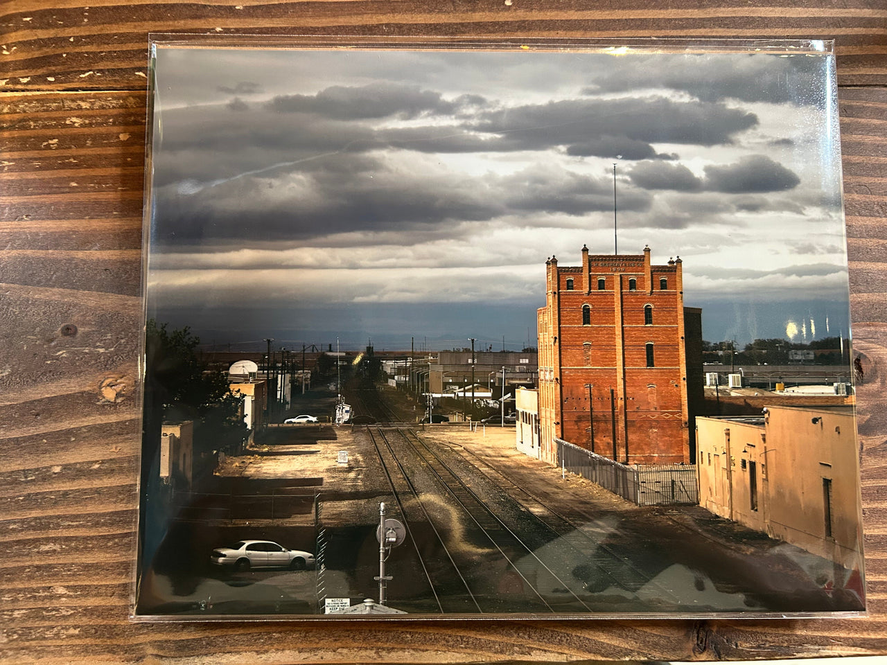 Vintage photograph of a cityscape with a tall brick building and train tracks, framed in glass on a wooden surface.